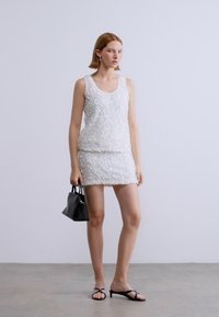 Λευκό αμάνικο τοπ με υφή και λεπτομέρειες που μοιάζουν με παγιέτες, σε συνδυασμό με μίνι φούστα που ταιριάζει. Το μοντέλο κρατάει μια μικρή μαύρη τσάντα.