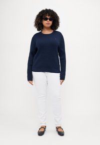 Mørkeblå strikket genser med et teksturert mønster, kombinert med hvite jeans og svarte flate sko. Solbriller fullfører looken.