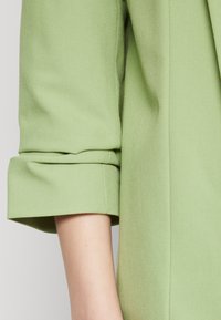 Ljusgrön, strukturerad tyg med rullad manschettdetalj, som visar upp rena linjer och en skräddarsydd framtoning. Närbild av ärmsektionen.