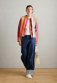 Gestreifte Jacke in mehreren Farben mit Rot-, Pink-, Gelb- und Blautönen, kombiniert mit einem weißen Oberteil und weit geschnittenen blauen Jeans, weißen Sneakers und einer beigefarbenen Tasche.