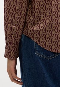 Manche d'une chemise marron à motifs avec des boutons au-dessus de la poche d'un jean en denim bleu et une main détendue sur le côté.