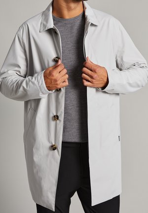 Hombre abotonándose un abrigo gris claro sobre suéter gris y pantalones negros contra un fondo liso.