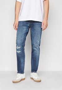 Jean en denim bleu clair avec une coupe slim, comportant des détails usés aux genoux. Associé à des baskets blanches et un t-shirt blanc uni.