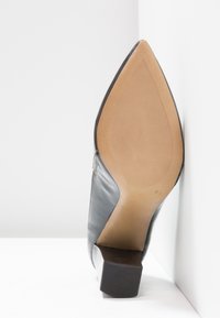 Chaussure à talon haut pointue pour femme noire vue de la semelle avec un fond beige texturé et un talon bloc.