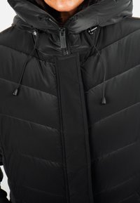 Veste rembourrée noire avec un col élevé et une capuche à cordon. Présente une fermeture éclair à l'avant et des motifs de couture verticaux. Texture lisse et légèrement brillante.