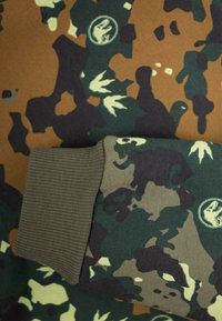 Sudadera de camuflaje multicolor con tonos de marrón, verde y negro, con un puño acanalado en verde oliva y logotipos. Textura de tela suave.