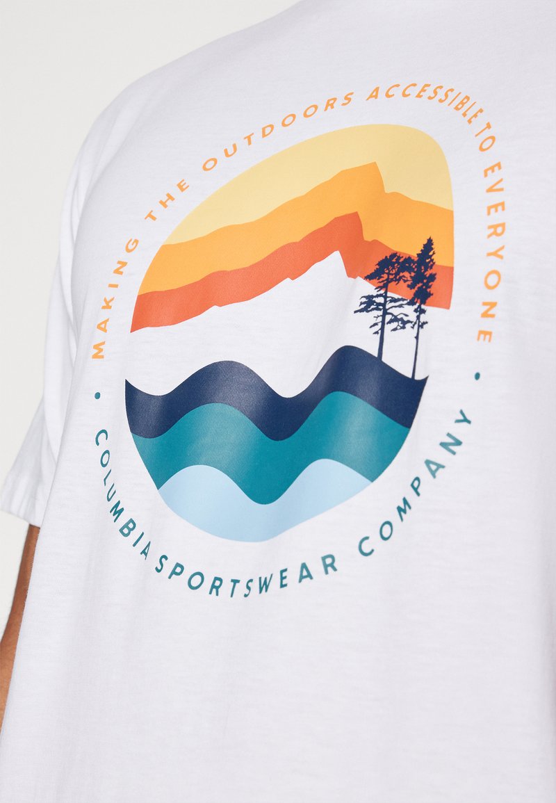Camiseta de algodón blanca con un gráfico circular que presenta montañas, árboles y olas en naranja, azul y verde azulado. El texto rodea el diseño.