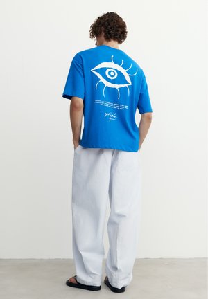 Tânăr care poartă un tricou albastru cu un grafic alb cu ochi și pantaloni albi largi, stând cu mâinile în buzunare pe un fundal simplu.