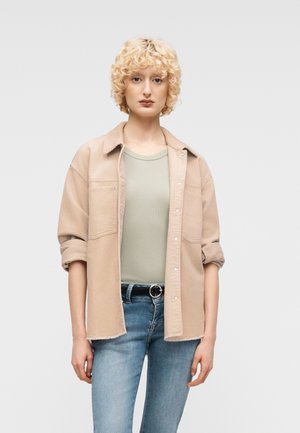 Young person with curly blonde hair wearing a beige button-up shirt over a light green top and blue jeans with a black belt.