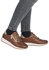 Person iført grå jeans og brune lædersneakers med guldfarvet lynlås og mønstrede snørebånd mod en hvid baggrund.