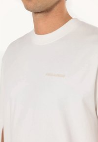 Hombre vistiendo una camiseta básica de cuello redondo blanca con un pequeño y sutil logo de texto beige en el pecho, sobre un fondo blanco.