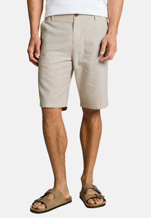 Man met beige knielange korte broek en bruine sandalen met gesp, staand tegen een effen witte achtergrond.