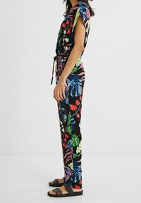 Blommig jumpsuit med korta ärmar; svart bakgrund; livfulla mönster i grönt, rött och blått; knuten midja; kombinerad med platta sandaler.