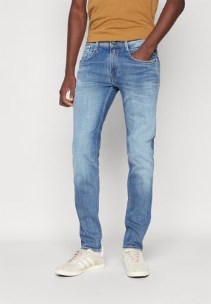 Mand iført lyseblå slim-fit jeans, sennepsskjorte og beige sneakers, stående med den ene hånd i lommen mod ensfarvet baggrund.