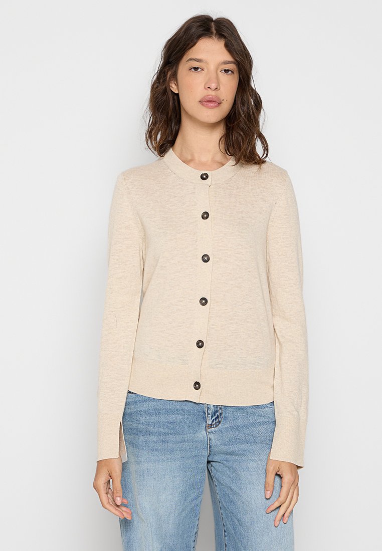 Kobieta z falującymi brązowymi włosami, ubrana w beżowy kardigan na guziki i jasnoniebieskie dżinsy, stojąca na tle gładkiej białej ściany.