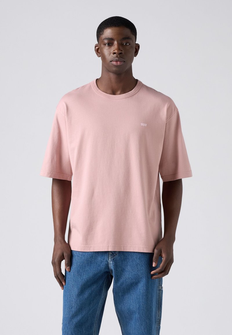 Homme portant un t-shirt ample rose clair uni et un jean bleu, debout devant un fond uni gris clair.