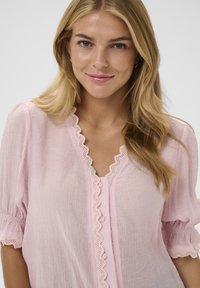 Blouse rose clair en tissu texturé, avec un bord en dentelle festonnée le long de l'encolure et du devant à boutons, et des manches bouffantes.