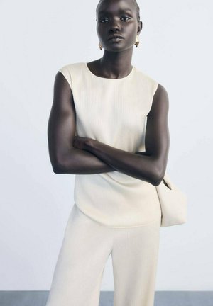 Light beige sleeveless top with a textured design, paired with flowing cream wide-leg trousers. Model stands with arms crossed, against a neutral background.