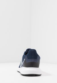 Marineblaue Adidas-Sneaker von hinten betrachtet, mit weißen Streifen, schwarzer Fersenschale und weißer Sohle auf einer weißen Oberfläche.