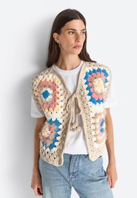 Crocheted vest in neutral beige with colorful circular patterns in pink, blue, yellow, and cream. Features a tie closure and fluffy accents.