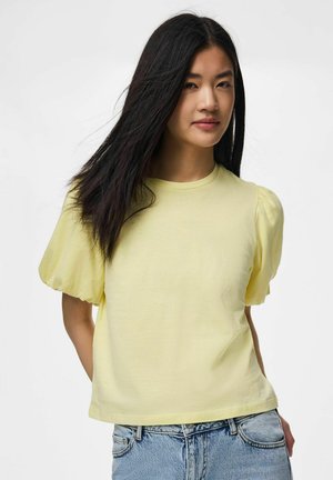Ung kvinne med langt mørkt hår iført en lys gul topp med pufferm er og blå jeans, stående mot en ensfarget hvit bakgrunn.