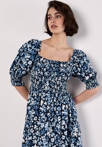 Robe fleurie avec un fond marine, présentant des manches bouffantes élastiquées, un décolleté carré et une taille froncée. Motif de fleurs bleues et blanches.