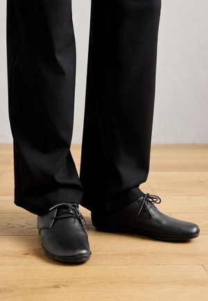 Chaussures habillées noires en cuir à lacets portées avec un pantalon noir sur un sol en bois clair.