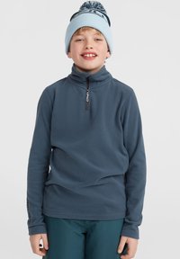 Fleece-tröja i mörkblått med kvartdragkedja och hög krage. Bärs med gröna byxor. Stickad bomullsbeanie med pom-pom på toppen.