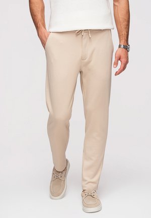 Homme portant un pantalon de jogging beige, des chaussures beiges à lacets, un haut en maille blanc et une montre-bracelet argentée, debout avec les mains détendues.