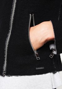 Svart ullblandad jacka med silverfärgade dragkedjor, med en sidoficka med en kontrasterande grå foder. Logotypen synlig nära nederkanten.