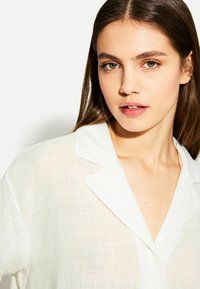 Camicia bianca a maniche lunghe con un tessuto testurizzato, dotata di colletto e vestibilità ampia. Il tessuto appare leggero e leggermente trasparente.