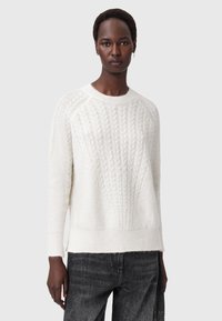 Persona che indossa un maglione bianco a trecce e jeans neri, in piedi contro uno sfondo grigio chiaro uniforme.