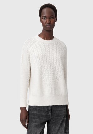Person, der einen weißen Kabelstrickpullover und schwarze Jeans trägt, steht vor einem einfarbigen hellgrauen Hintergrund.