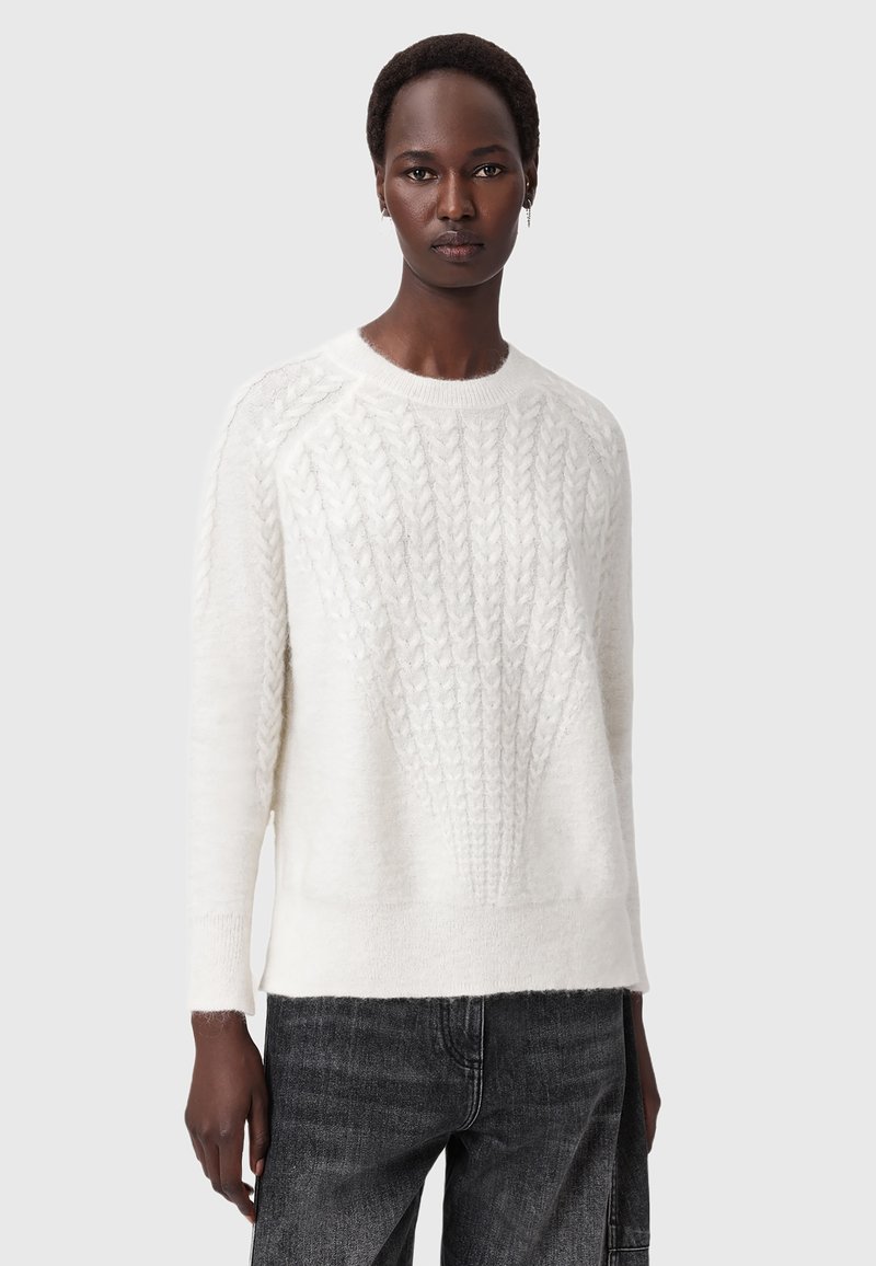 Persona che indossa un maglione bianco a trecce e jeans neri, in piedi contro uno sfondo grigio chiaro uniforme.
