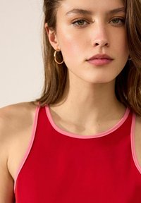Röd ärmlös topp med rosa kant, featuring en rundad halsringning och en slät textur. Accentuerande hoops örhängen fulländar looken.