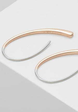 Deux fines boucles d'oreilles hoop ouvertes avec la moitié supérieure en or rose et la moitié inférieure en argent, reposant sur une surface blanche.
