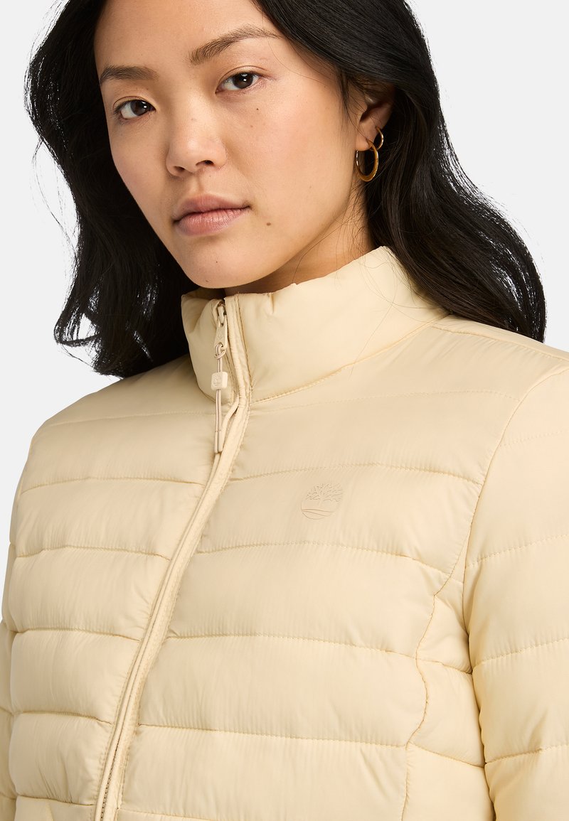 Giacca puffa beige con colletto alto, caratterizzata da un design trapuntato, zip frontale e un piccolo logo in rilievo sul petto.