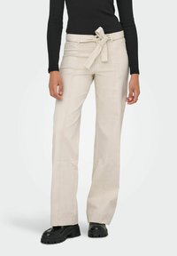 Femme portant un pantalon beige taille haute à jambes larges avec une ceinture nouée, un haut noir à manches longues et des chaussures noires épaisses sur fond blanc.
