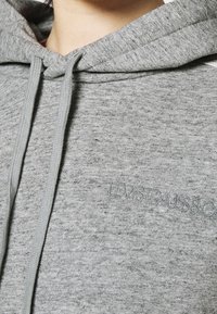 Un hoodie gris en tissu doux et texturé avec une capuche à cordon. Présente un logo discret en gris foncé sur le devant.
