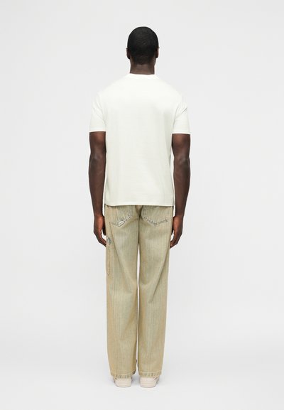 Άνδρας που φοράει απλό λευκό κοντομάνικο μπλουζάκι, μπεζ άνετο τζιν και λευκά αθλητικά παπούτσια, όρθιος με την πλάτη προς το μέρος του θεατή, μπροστά σε έναν απλό φόντο.