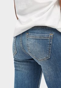 Slim fit modré džíny z denimového materiálu s mírným vyblednutím, které mají dvě zadní kapsy a viditelné šití. Textura vypadá hladce a pevně.