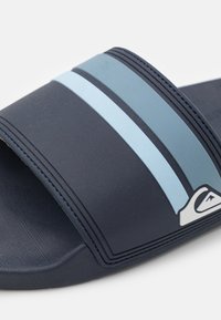 Tamno plava natikača ima tri horizontalne svijetloplave pruge i teksturiranu unutrašnjost. Izrađena je od gume s naglaskom na logotip.