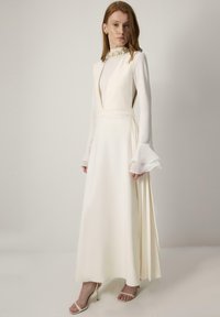 Lang witte jurk met een diepe V-hals, lange doorschijnende belmouwen en een getailleerde taille. Kenmerkt zich door een hoge kraag met parelaccenten en een vloeiende rok.