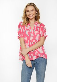 Pink bluse med abstrakte blå og lyserøde m�ønstre, korte puffærmer, V-hals og en flagrende kant. Parret med blå jeans.