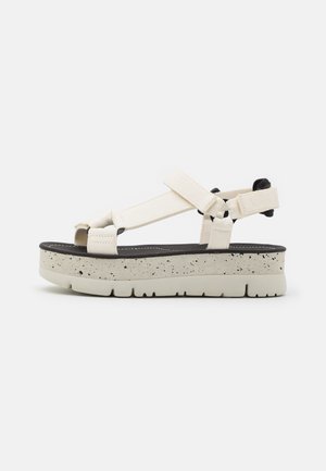 Platform sandal with cream elastic straps, black footbed, and a speckled gray sole. Features adjustable straps for a secure fit.