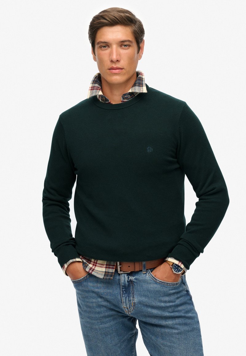 Jersey de color verde oscuro con textura acanalada, superpuesto sobre una camisa de cuadros. Presenta un escote redondo y un logo bordado en el pecho.