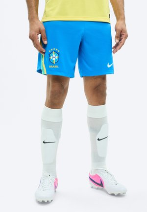 Jucător de fotbal masculin purtând pantaloni scurți albastri cu logo-ul Braziliei, apărători de tibie albi Nike, și ghete albe cu accente roz și negre.