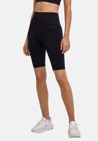 Femme portant un short de vélo noir taille haute et des chaussures de course blanches, se tenant avec les bras détendus le long du corps sur un fond uni.