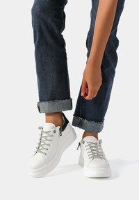 Sneakers bianchi in pelle con lacci argentati, dettagli neri sul tallone e una spessa suola bianca. Indossati con jeans in denim scuro con risvolto.