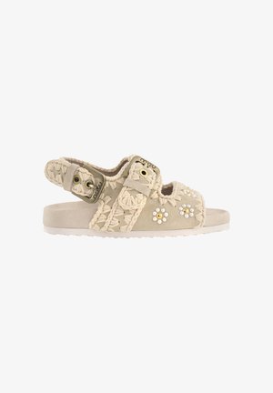 Beige åben-tå sandal med hæklede remme, justerbar spænde og små perle- og guldbelagte blomsterudsmykninger på siden.